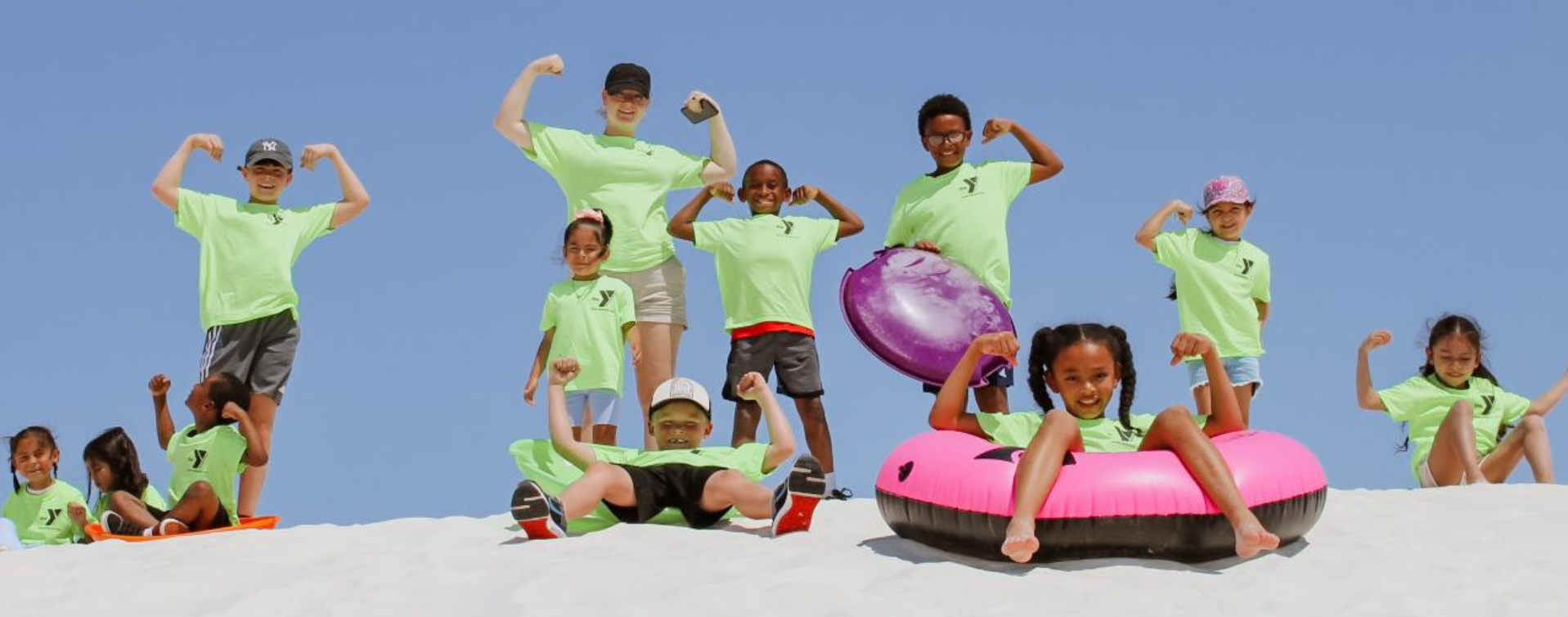 A diverse group of children wearing bright green ASYMCA shirts pose on sand dunes under a clear blue sky, smiling and flexing their arms during a summer camp activity.
