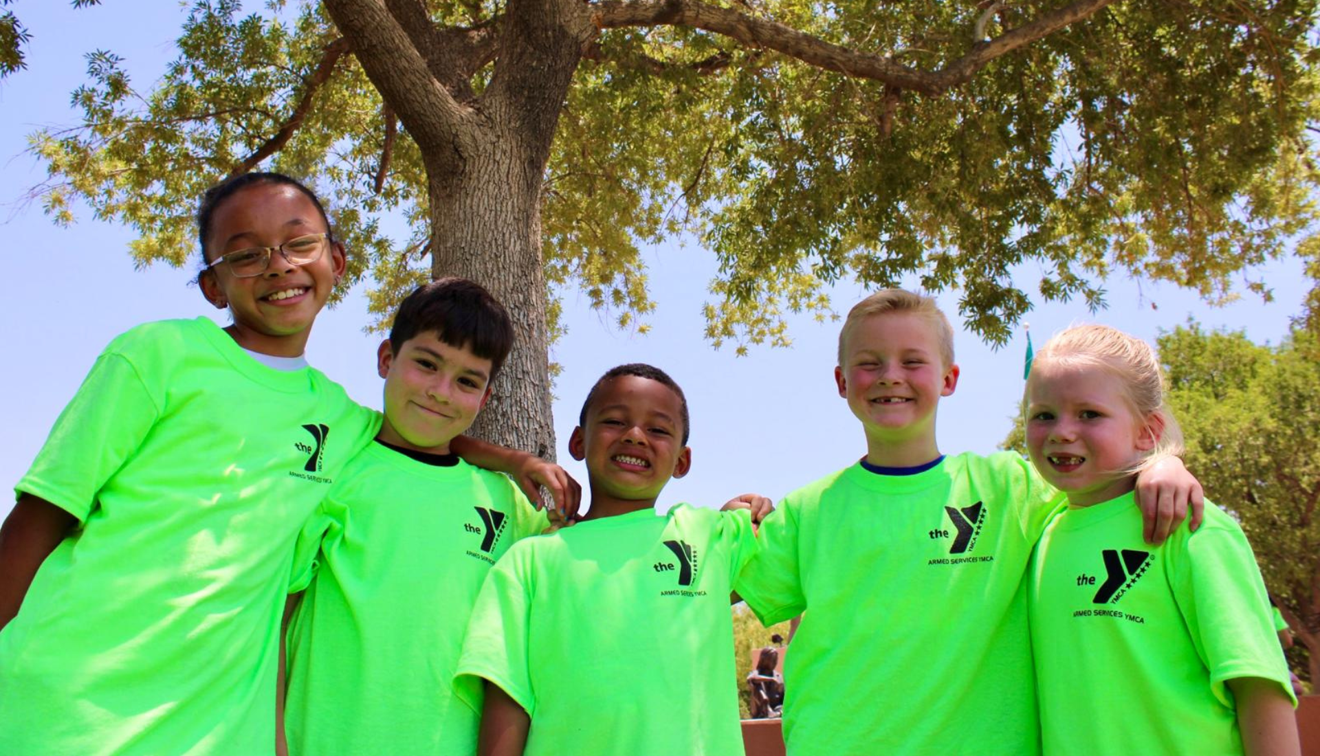 Children wearing bright green Armed Services YMCA shirts stand together outdoors, smiling during a camp activity.