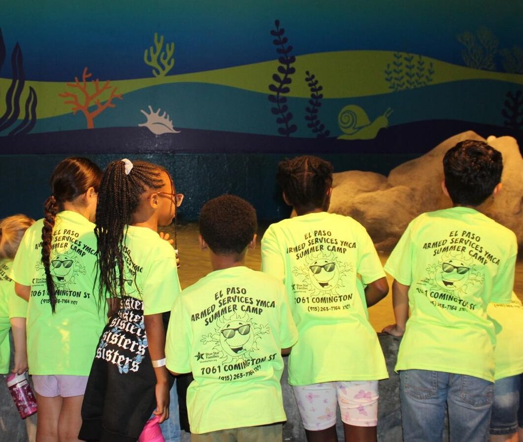 Children in neon green Armed Services YMCA El Paso Summer Camp shirts looking at an underwater-themed exhibit during a field trip.