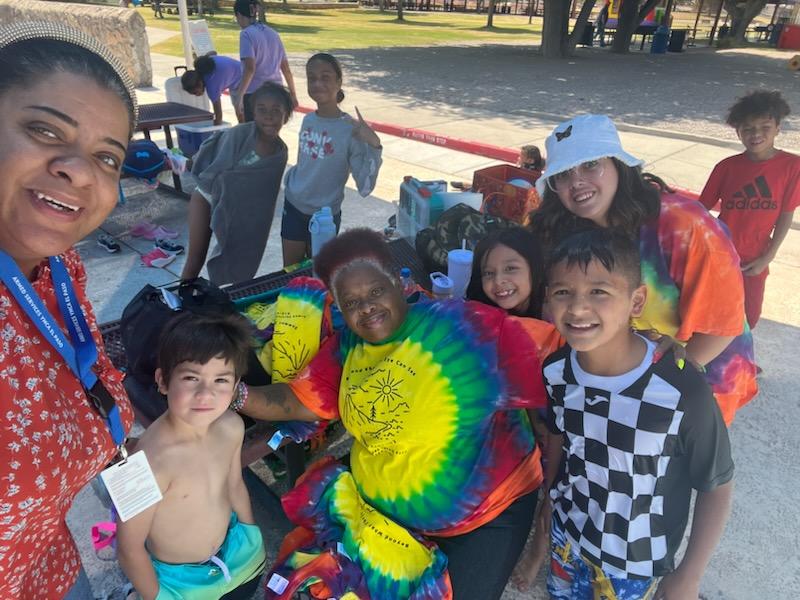 Operation Summer Camp staff and children smiling together at a splash pad and park during a summer outing.